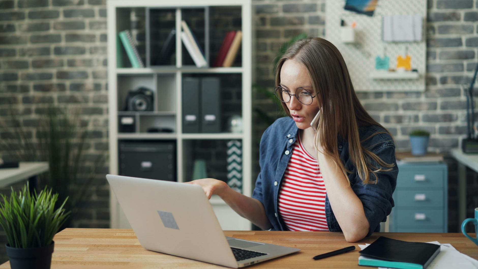 Focused young woman working on laptop while talking on a phone in a modern office.