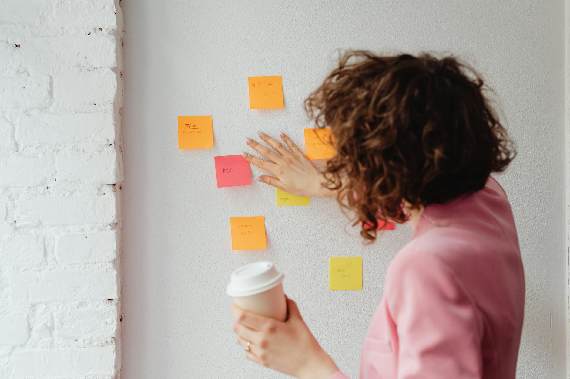 A woman in a pink blazer organizing sticky notes on a white wall while holding a coffee cup.